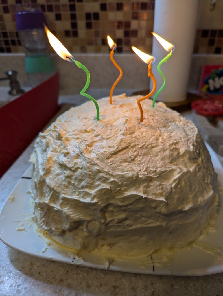 A large rounded cake, iced white, with four thin, curly, lit candles.