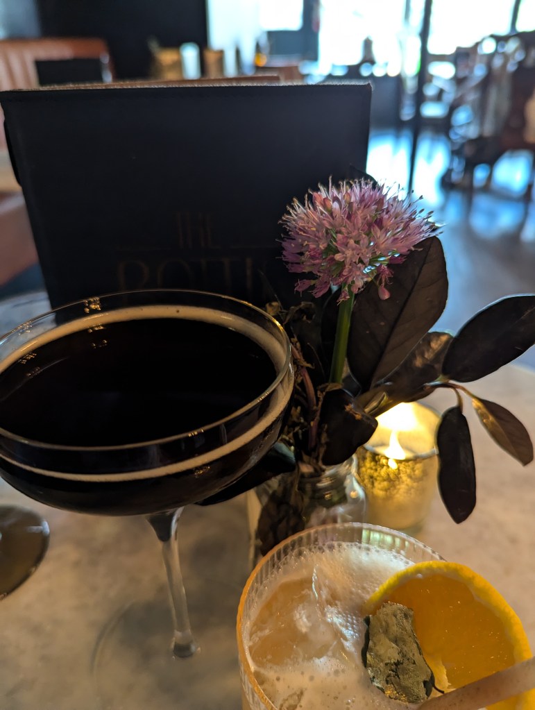 Two cocktails arranged around a small floral centerpiece, a candle, and a leatherbound menu