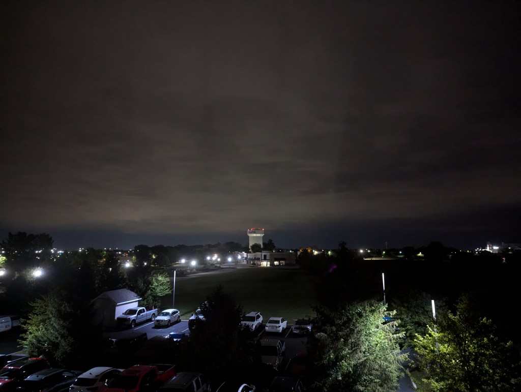 A night sky overlooking a parking lot and a distant water tower, taken from a hotel window